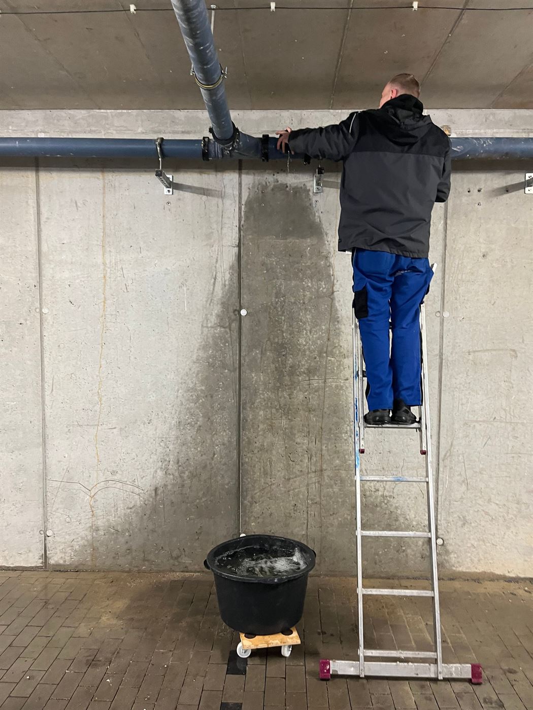 Mitarbeiter auf Leiter arbeitet an einem Fallrohr in einer Tiefgarage